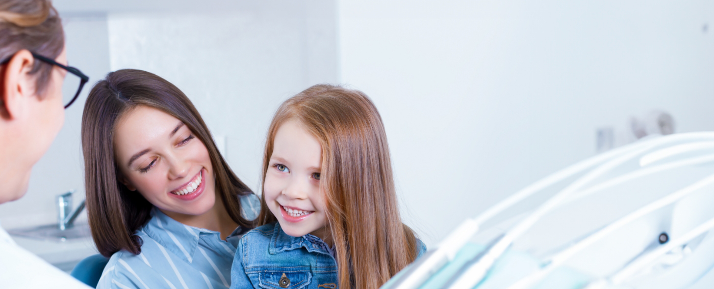 Young girl in her mothers lap grinning at her pediatric dentist