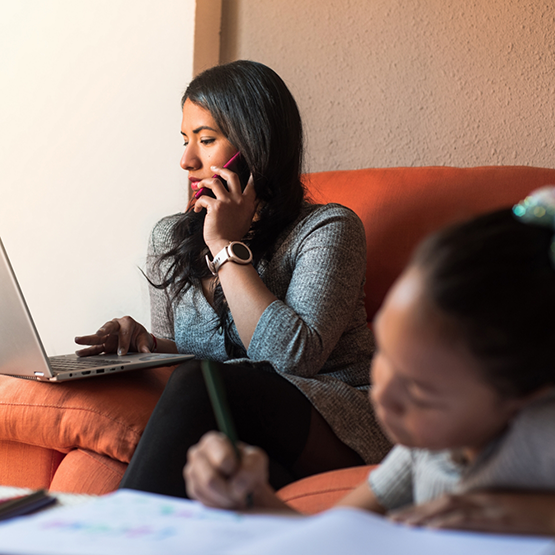 Mother talking on phone and looking at laptop on couch while daughter colors in foreground